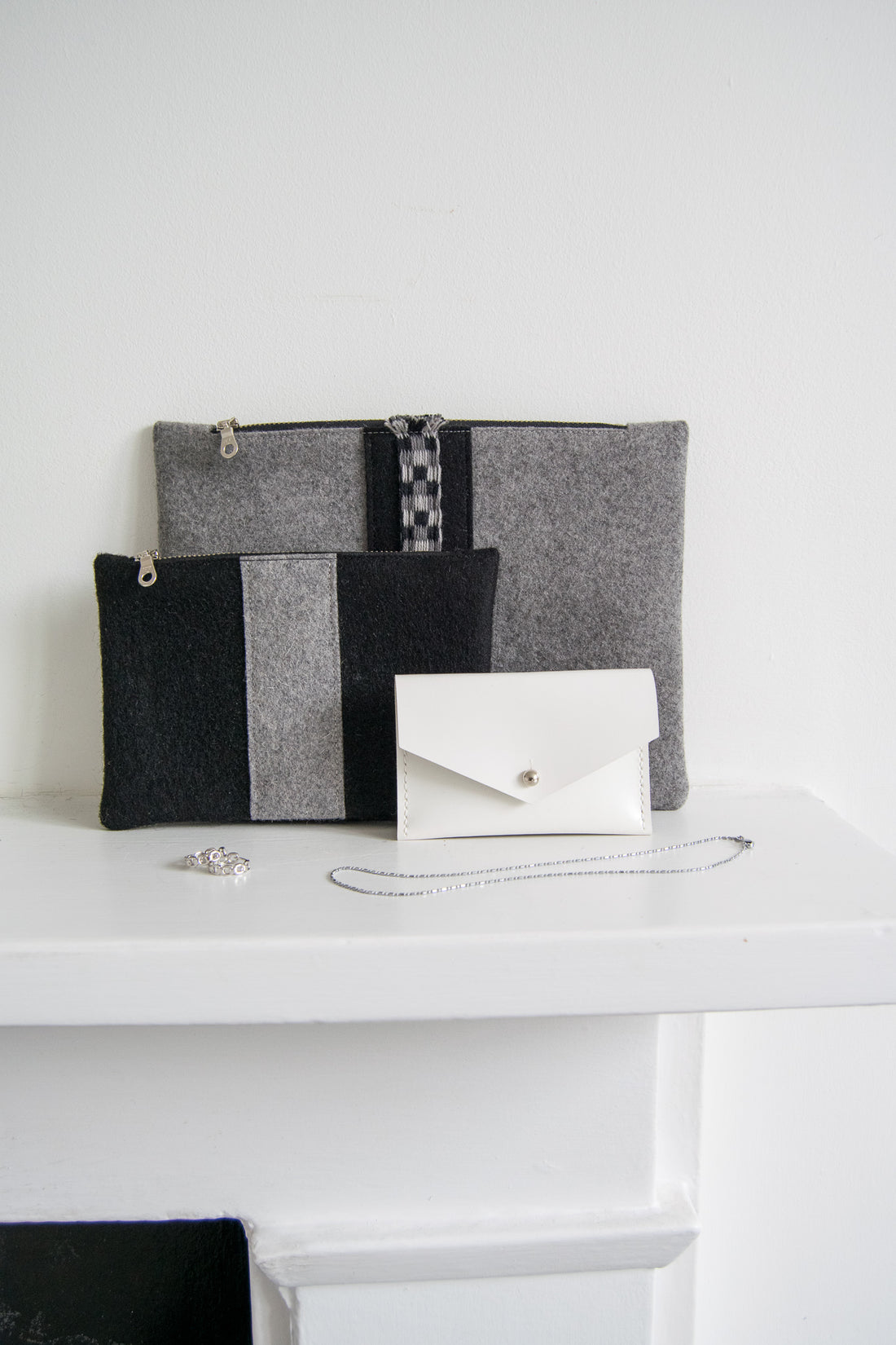 Gray and black wool zip pouches paired with a white leather card holder and silver jewelry on white mantel.