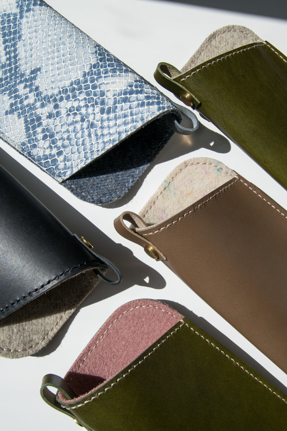 A flatlay of 5 leather sunglasses sleeves in blue, olive green, black and light brown leather.